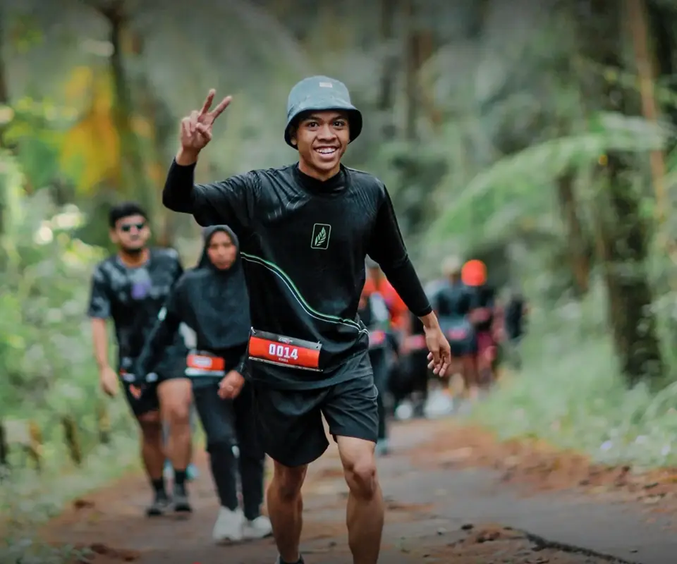 Pelari tersenyum mengacungkan dua jari di jalur hutan, diikuti pelari lain